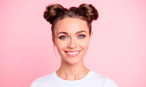 young woman with hair in space buns smiling against pink background